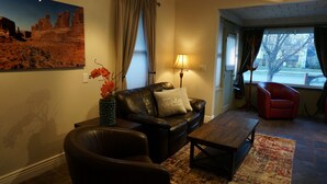 A living room with a leather sofa, a coffee table, a red armchair, a lamp, and a painting of a desert landscape.