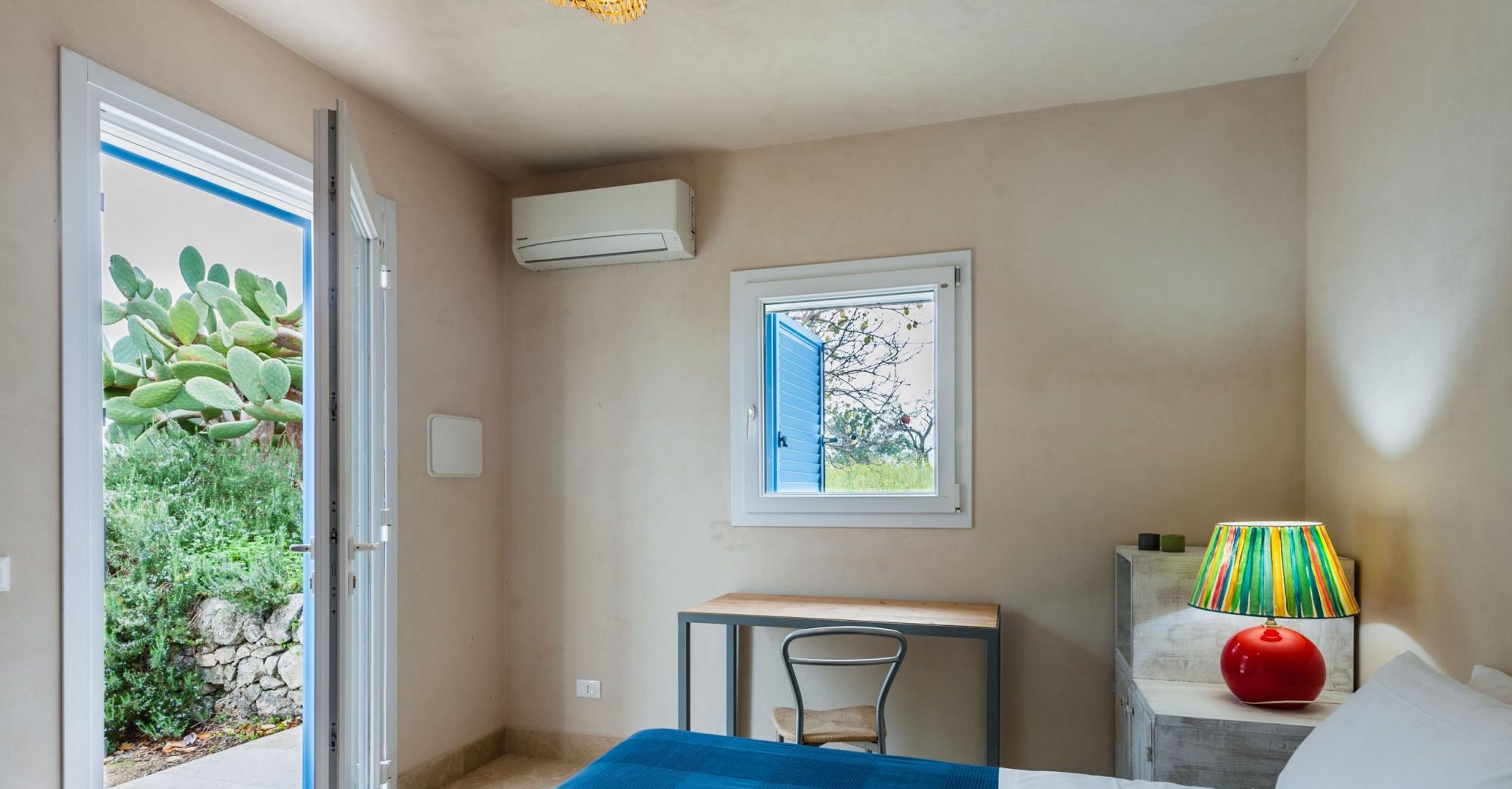 A room with a bed, a desk, a chair, a window with a view of greenery, an air conditioning unit, and a colorful lamp.