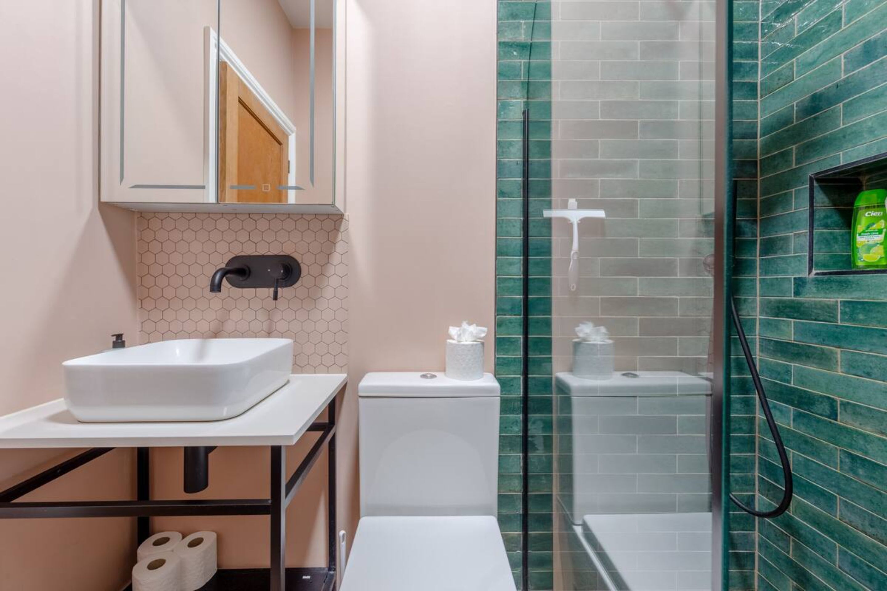 A bathroom with a white sink, toilet, and shower. There is a mirror above the sink and a shelf with toiletries.