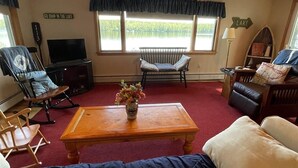 A cozy cabin interior with a wooden ceiling, a large window overlooking a lake, a television, and a fireplace. - Tremont Limit 10 4 Bedroom Villa by RedAwning (Bridgton)