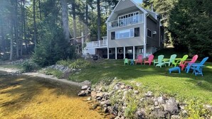A two-story house with a deck, surrounded by trees and a clear stream. - Tremont Limit 10 4 Bedroom Villa by RedAwning (Bridgton)