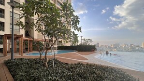 A rooftop pool area with a view of the city skyline, surrounded by greenery and a wooden deck. - Supertramp House - Gold View Apartment (Ho Chi Minh City)