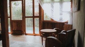 A room with a wooden floor, a round table, and two chairs. There are large windows with curtains, and a view of greenery outside. - Sarini Villas (Sukawati)