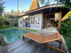 Eine Holzterrasse mit Pool, Liegestuhl und einem Haus mit großen Fenstern und einem Strohdach.