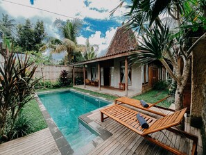 Ein Poolbereich mit Holzliegen, einer überdachten Terrasse und üppiger tropischer Vegetation.