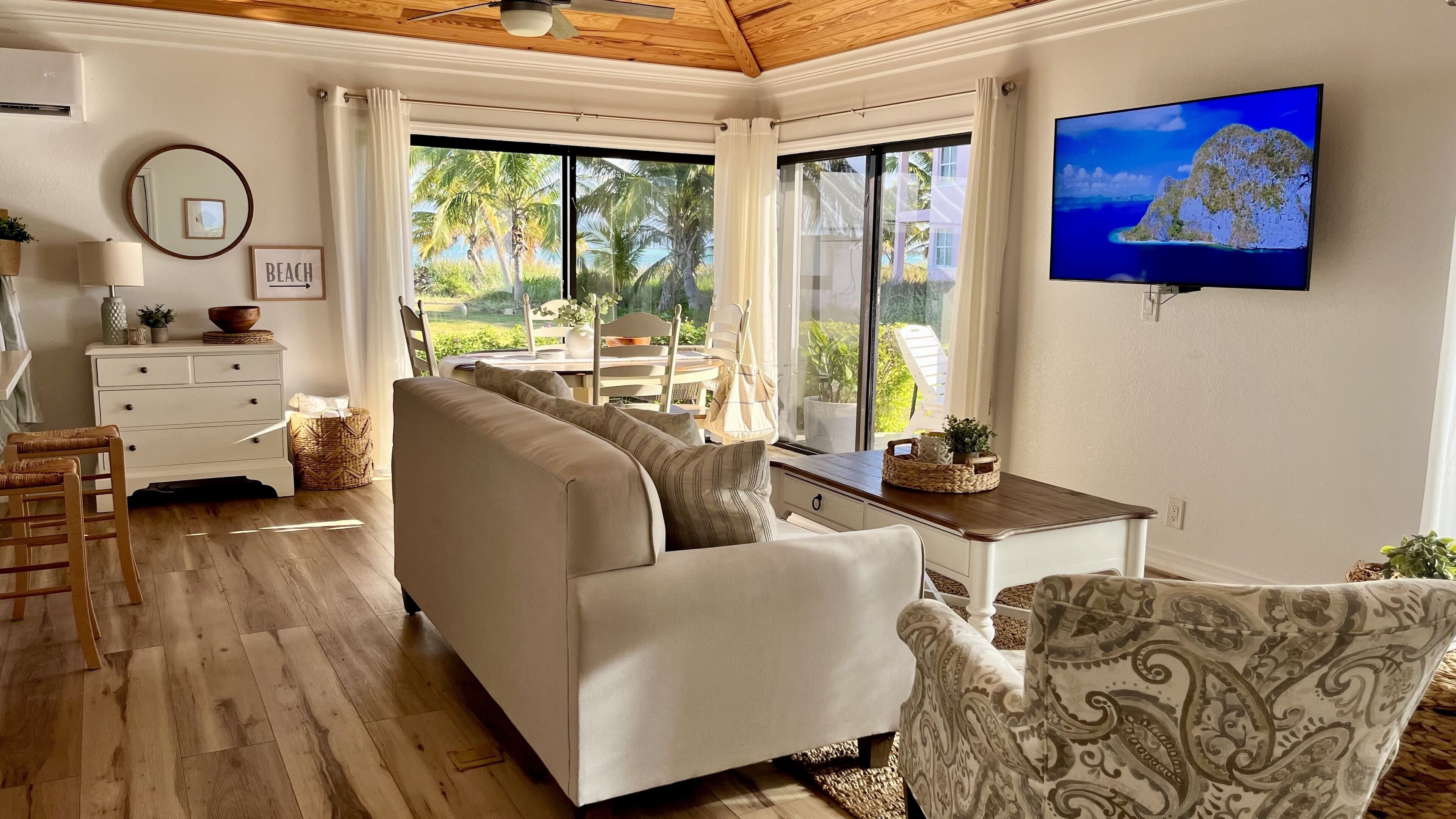 A spacious living room with a beige sofa, a patterned armchair, a wooden coffee table, and a television mounted on the wall.