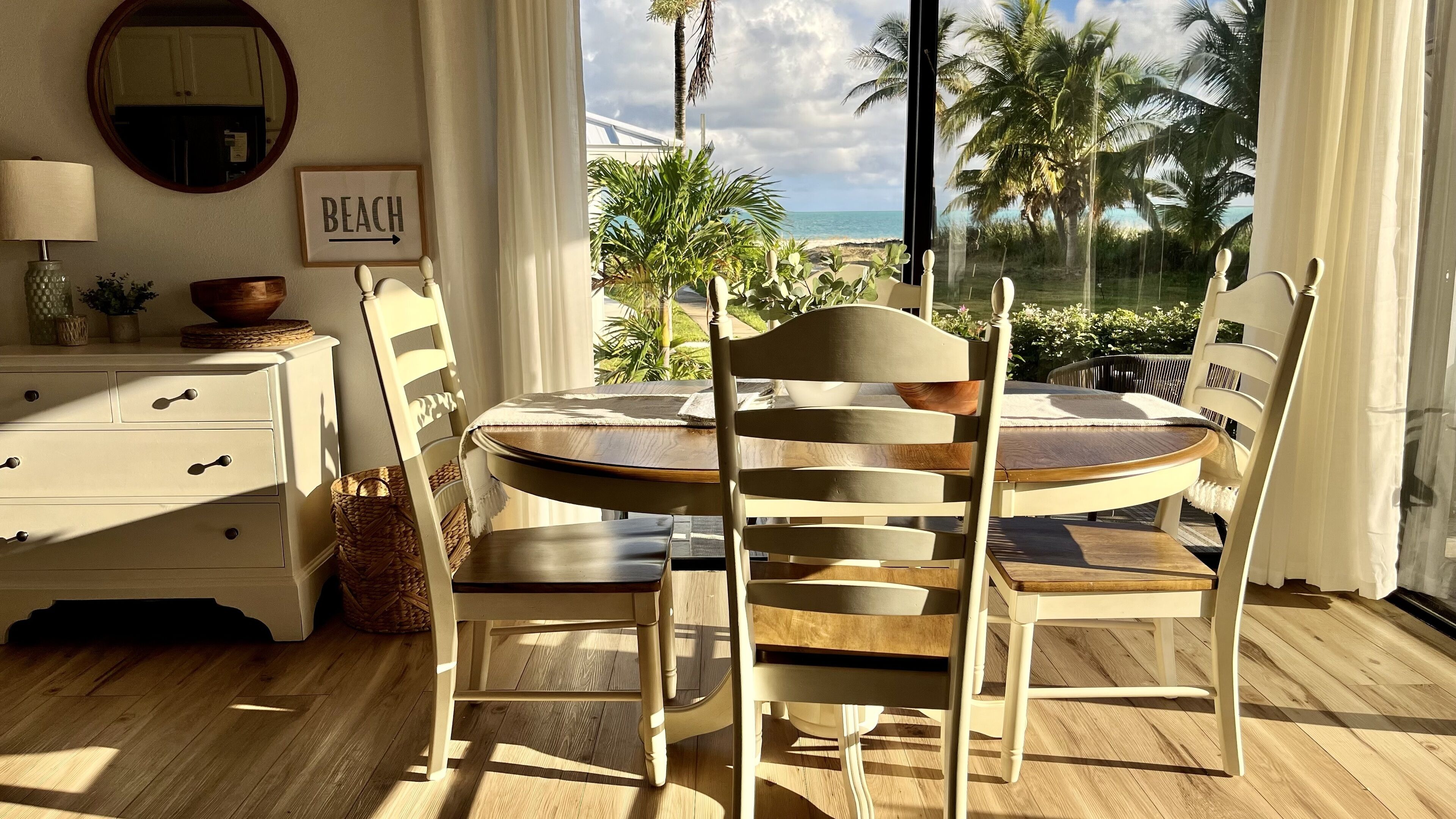 A dining area with a round table and chairs, a white dresser, a mirror, and a sign that says "BEACH".