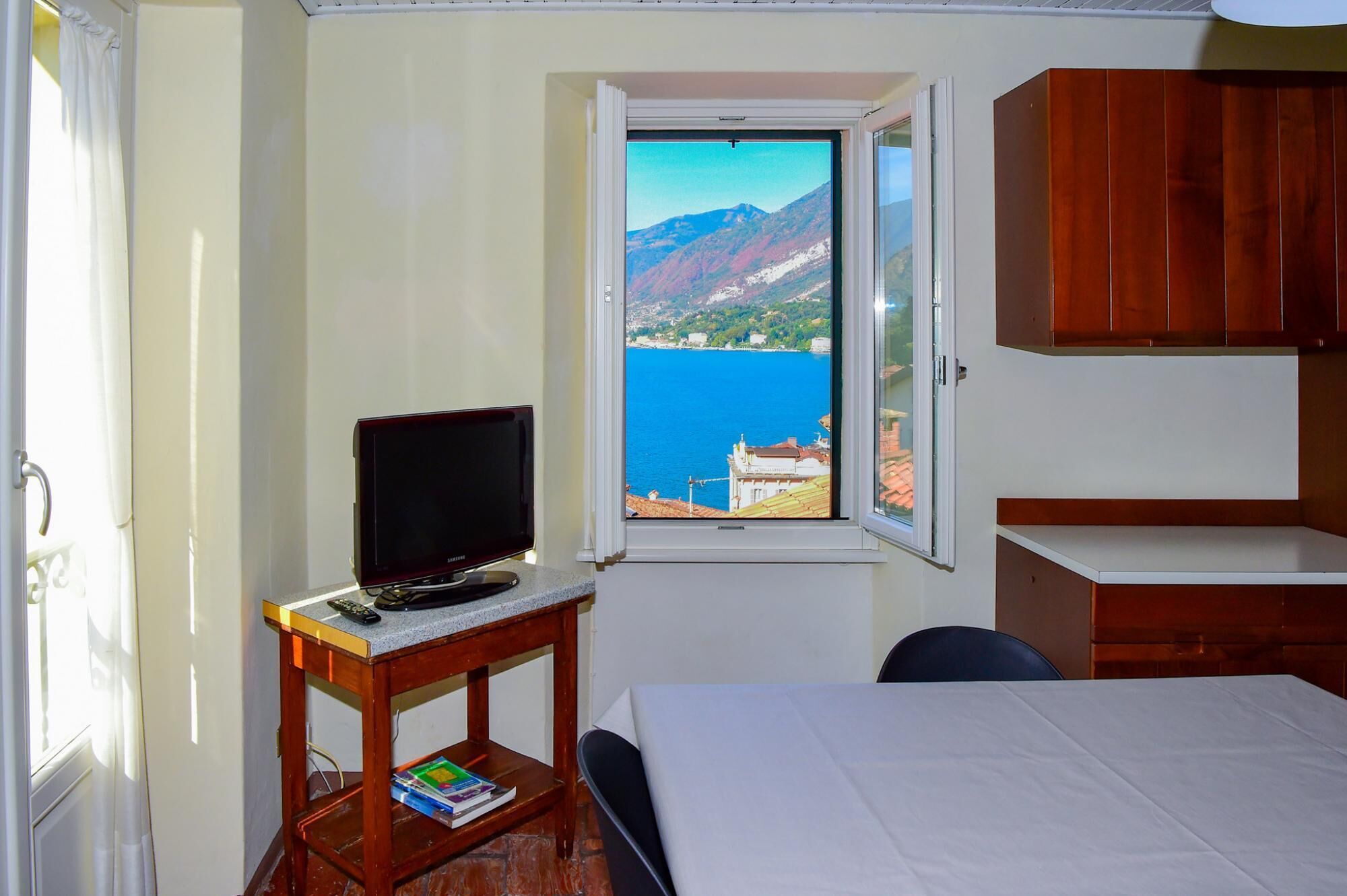 Quarto com cama, escrivaninha com televisão, janela com vista para montanhas e mar, e armários de madeira.