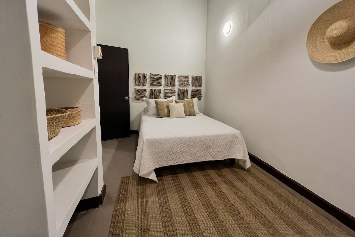 Une petite chambre à coucher, bien rangée, avec un lit, une étagère avec des paniers, une lampe fixée au mur et un chapeau de paille.