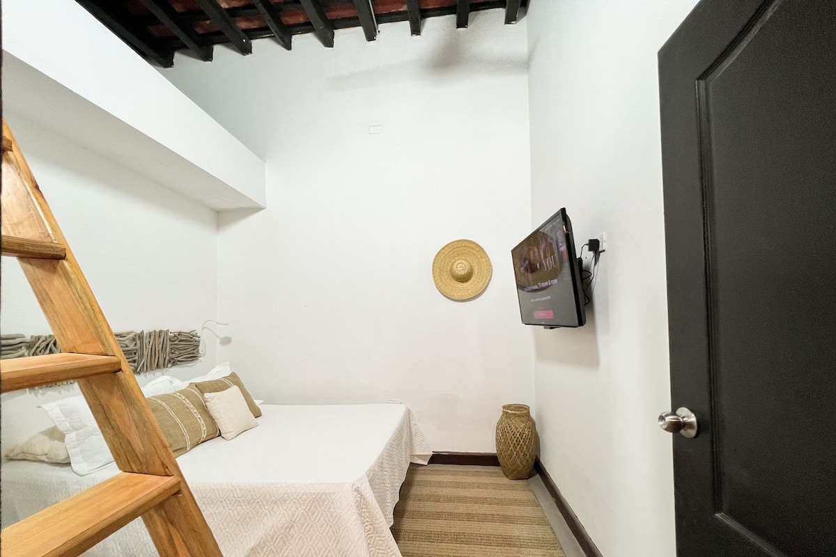 Une chambre avec un lit, une échelle en bois, un panier en osier et un téléviseur fixé au mur.