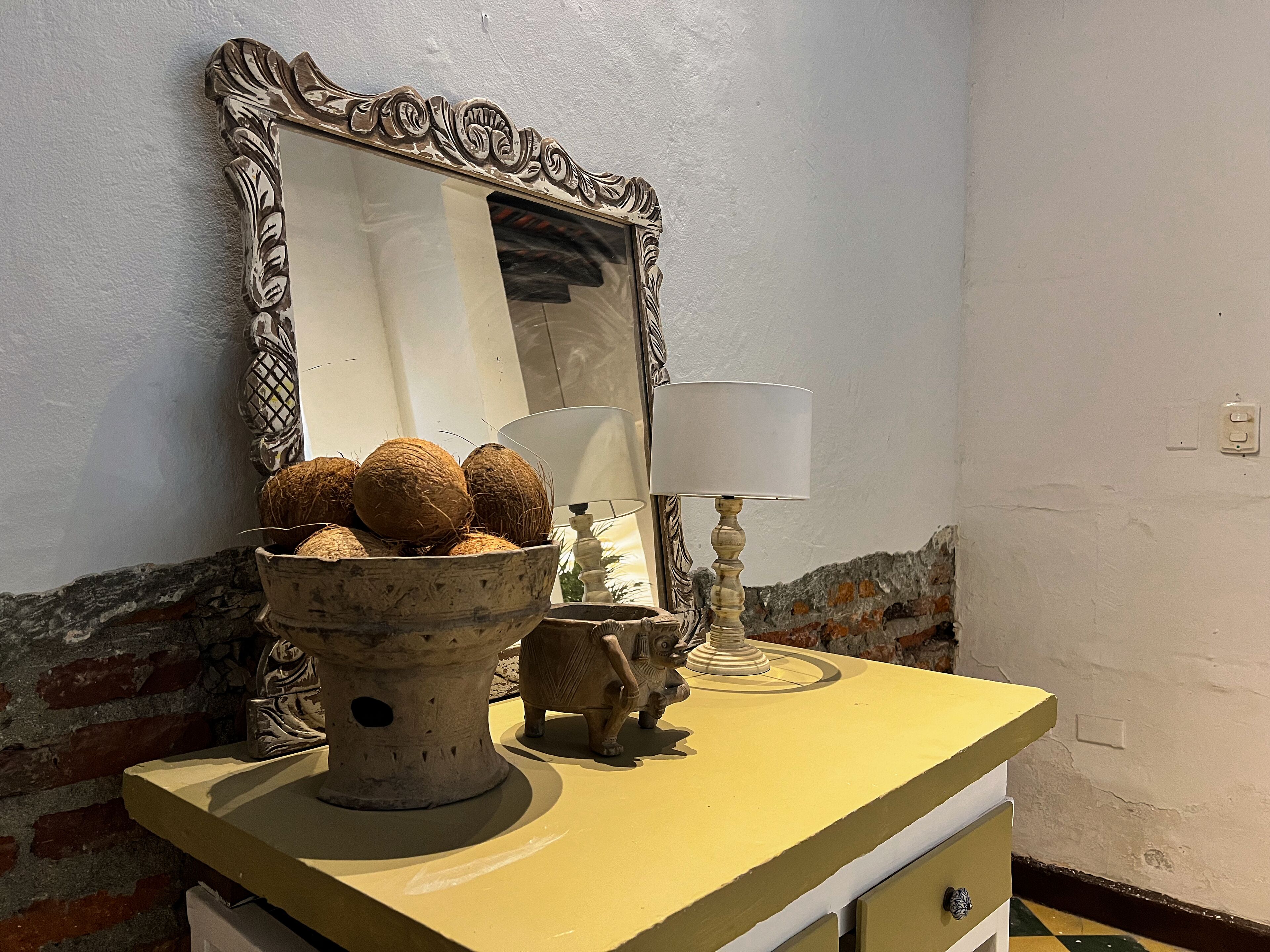 Een badkamer met een geel wastafelmeubilair, een decoratieve spiegel met een sierlijk frame, een schaal kokosnoten, een klein beeldje en een lamp.