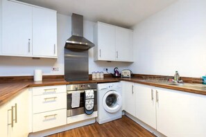 A compact kitchen with white cabinets, a stainless steel range hood, a built-in oven, a washing machine, and a wooden countertop.