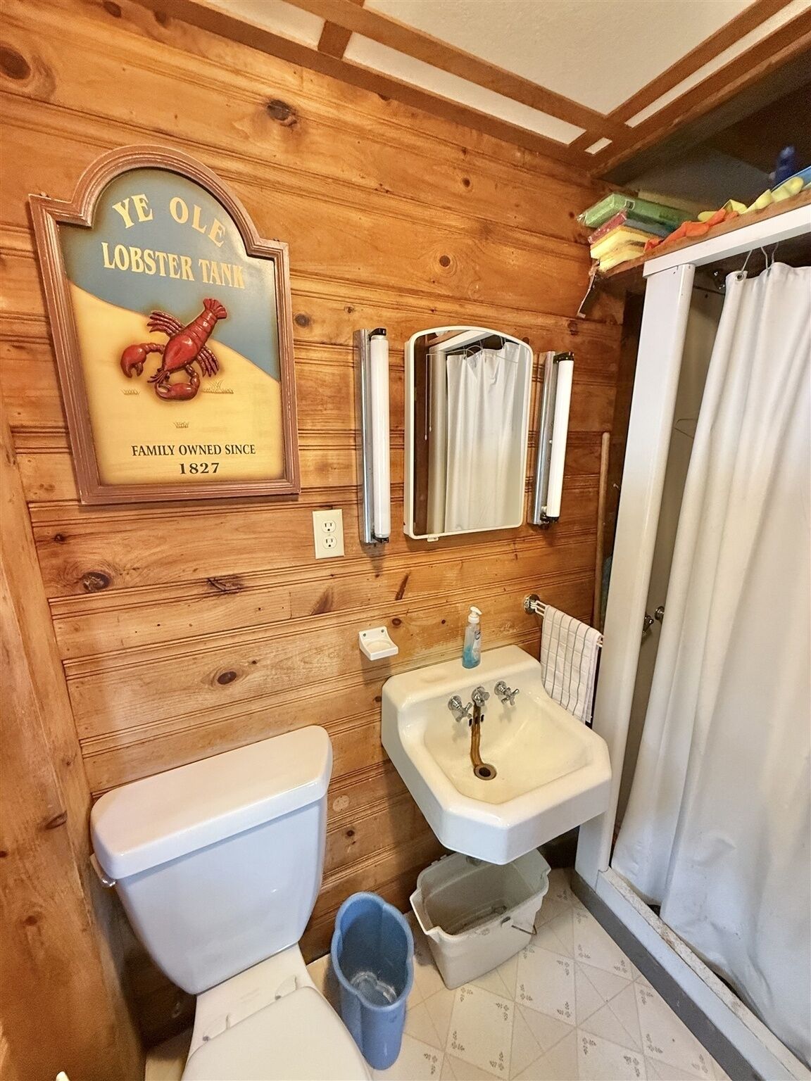 A bathroom with wooden walls, a toilet, a sink, and a shower curtain. A sign above the sink reads "YE OLE LOBSTER TANK FAMILY OWNED SINCE 1827".