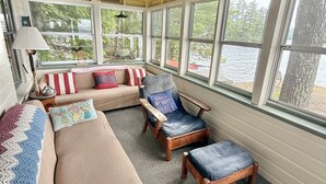 Ein gemütliches Zimmer mit Holzbalkendecke, großen Fenstern, einer Couch, einem Sessel und einer Lampe.