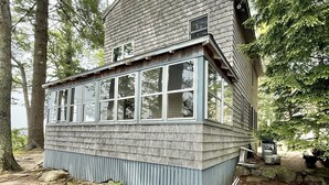 Ein zweistöckiges Holzhaus mit einer breiten Veranda mit Fenstern, umgeben von Bäumen und einer naturnahen Umgebung.