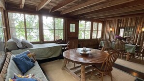 A rustic cabin interior with a wooden dining table, chairs, a sofa with cushions, and large windows offering a view of the outdoors. - Mackinney Camp - Limit 8 4 Bedroom Cottage by RedAwning (Standish)
