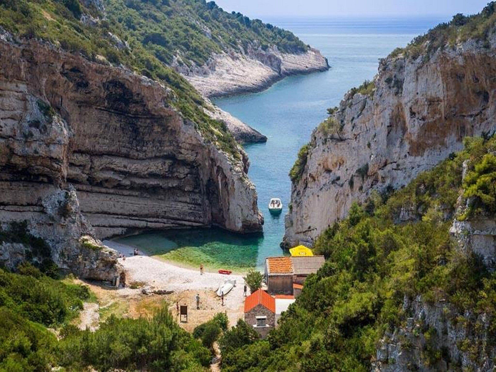 Paisaje costero con una estrecha vía fluvial, acantilados rocosos y una pequeña playa con un edificio y un bote.