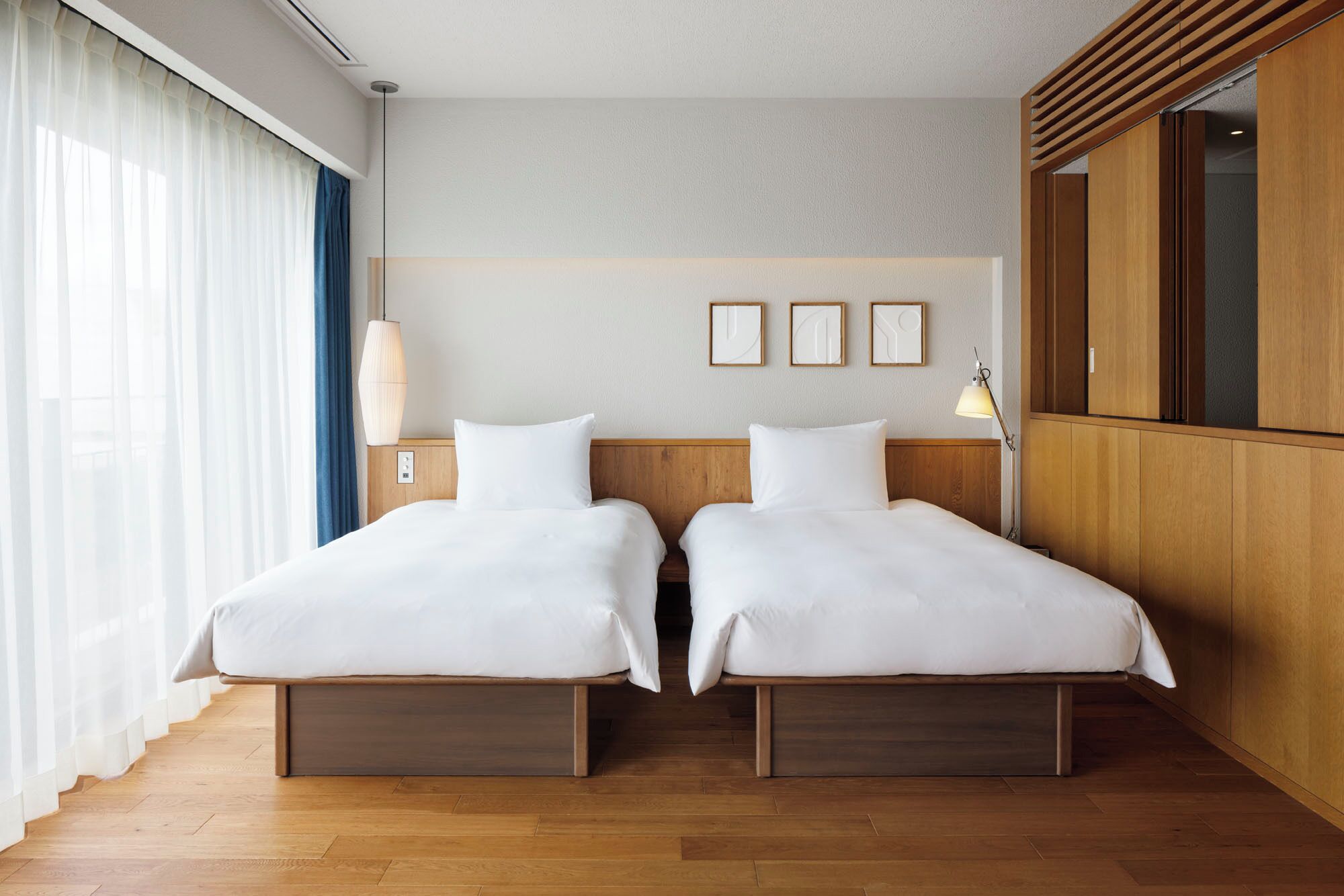 A hotel room with two beds, a large window with sheer curtains, a wooden headboard, and a wall-mounted TV.