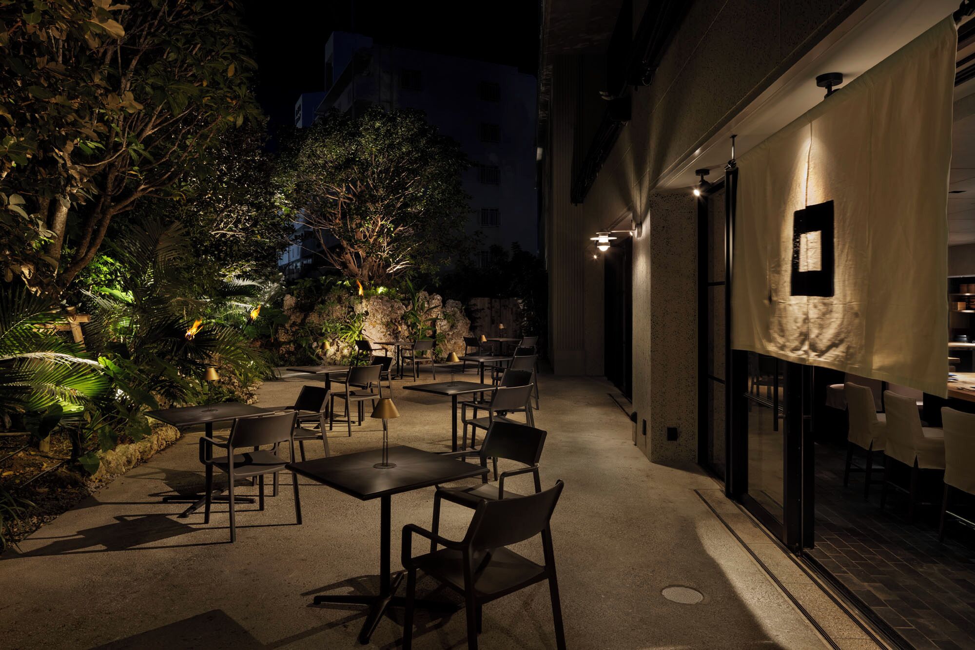 Un espace de restauration en plein air, avec des tables et des chaises, entouré d’une végétation luxuriante et bordé par la façade d’un bâtiment.