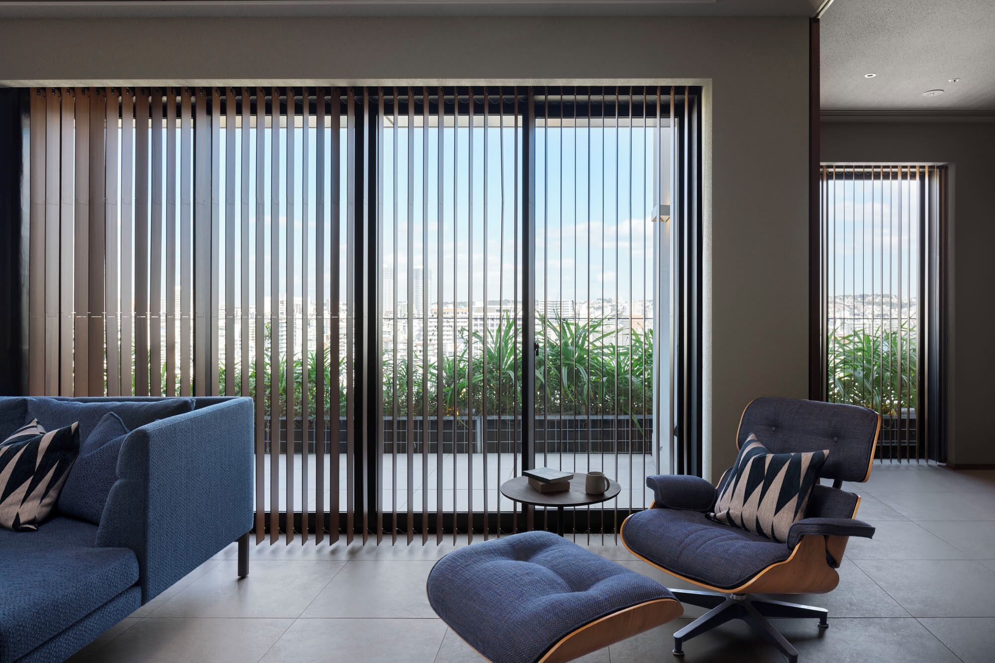 A modern living room with a sofa, armchair, and ottoman, large sliding glass doors, and a view of a cityscape.