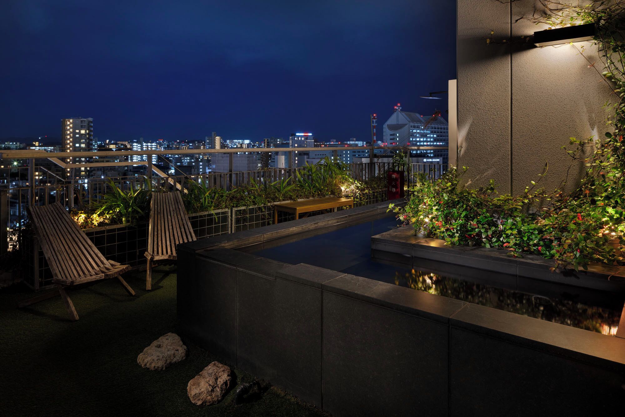 A rooftop terrace with a wooden bench, potted plants, and a view of the cityscape at night.