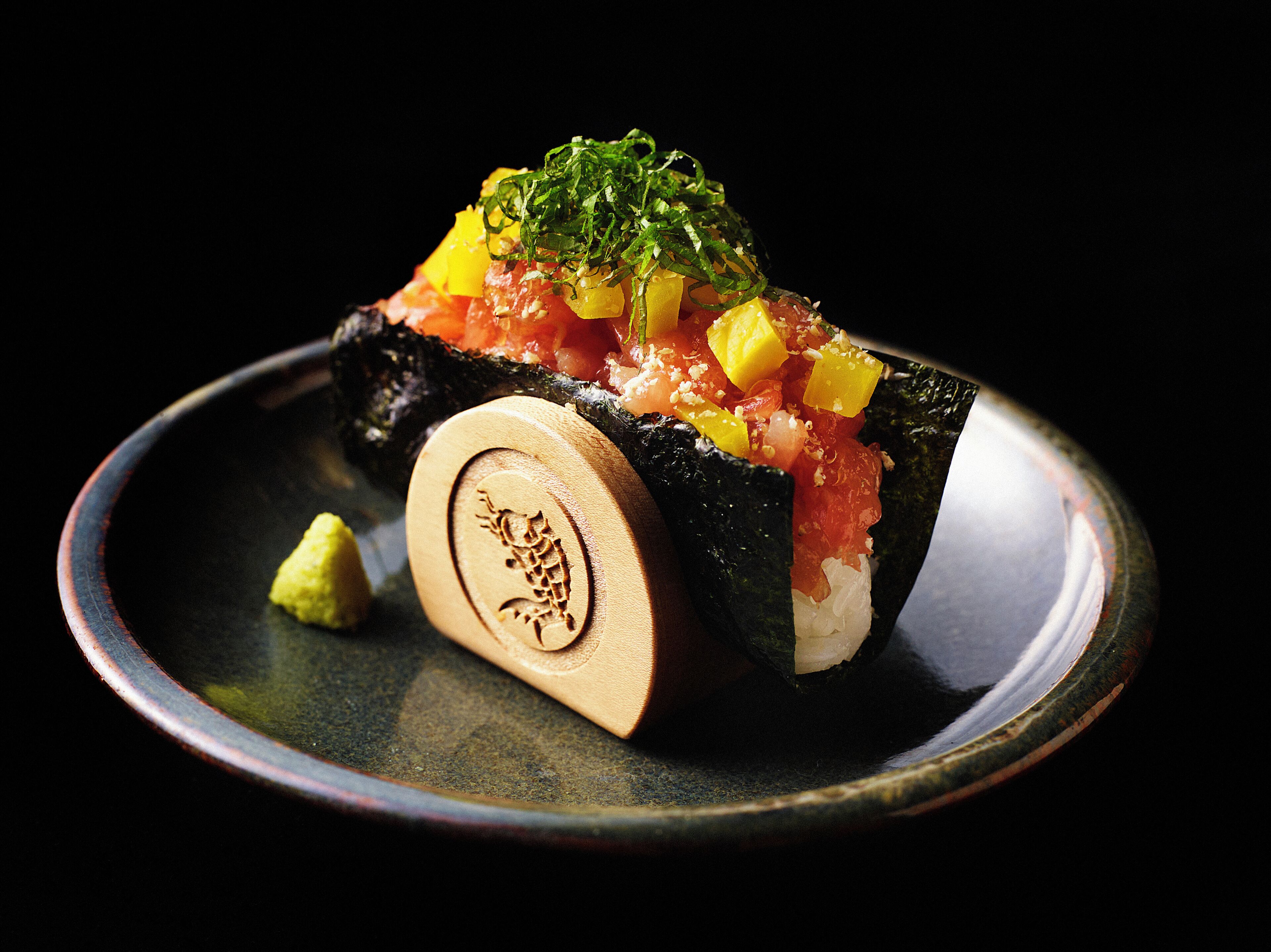 Un plat de sushis, accompagné d’une spatule à sushi en bois, présenté sur une assiette sombre.