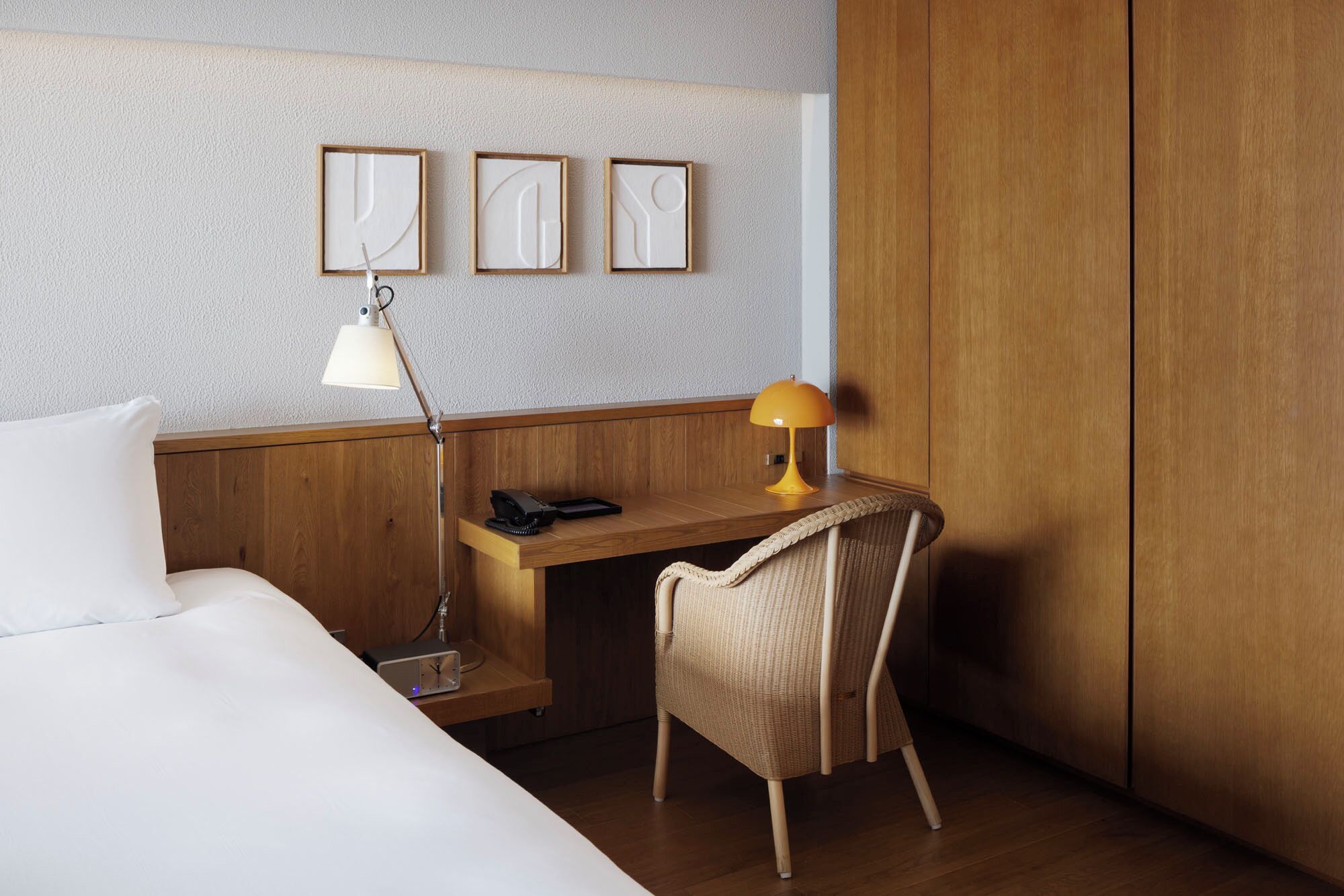 Quarto de hotel com cama, escrivaninha com abajur, cadeira e três quadros na parede.