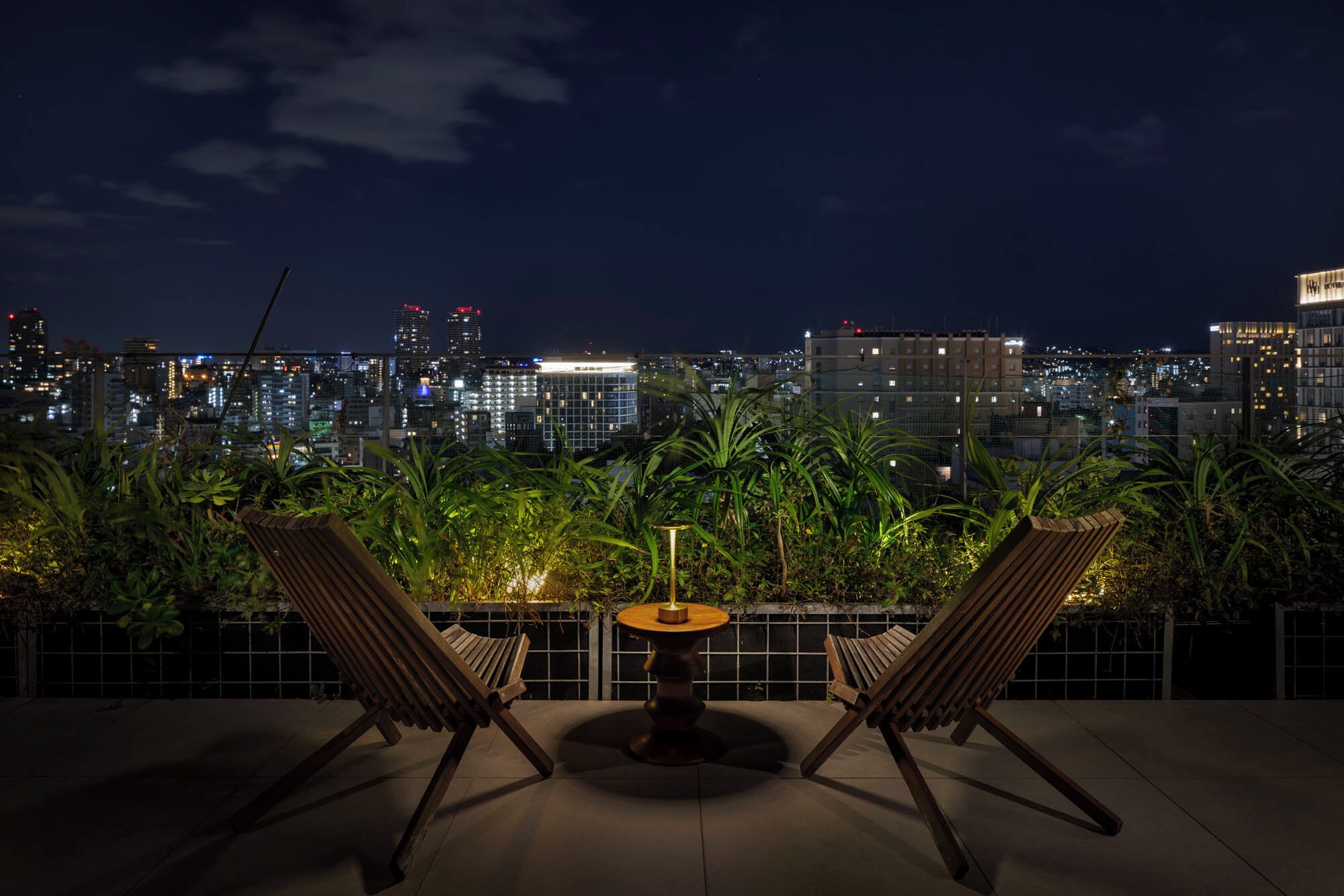 A rooftop terrace with two wooden lounge chairs and a small round table, overlooking a cityscape at night.