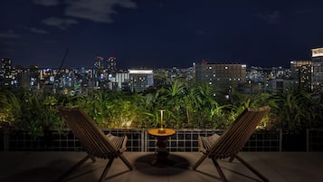 Une terrasse sur le toit, avec deux fauteuils de jardin en bois et une petite table ronde, d’où l’on peut admirer la vue sur la ville illuminée la nuit.