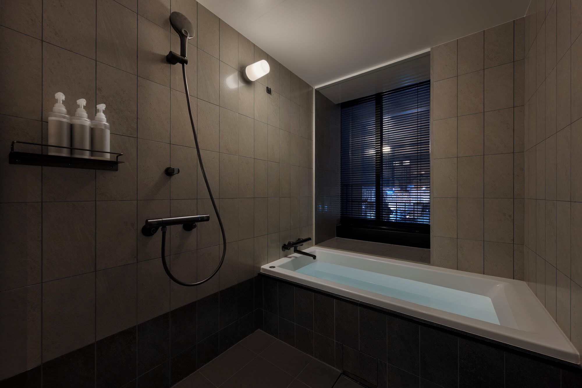 A modern bathroom with a bathtub, a handheld showerhead, and a window with blinds.