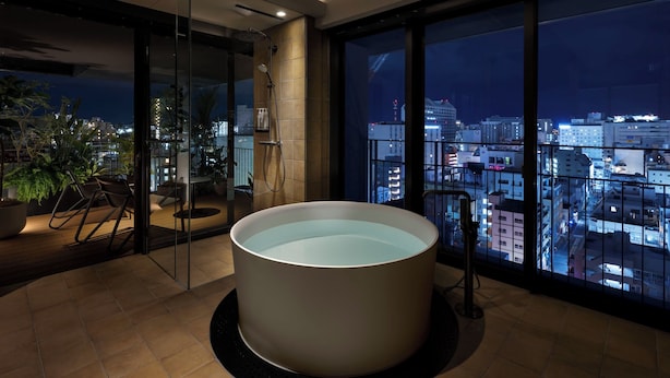 A modern bathroom with a large bathtub, a glass shower enclosure, and a city view through large windows. - Southwest Grand Hotel (Naha)
