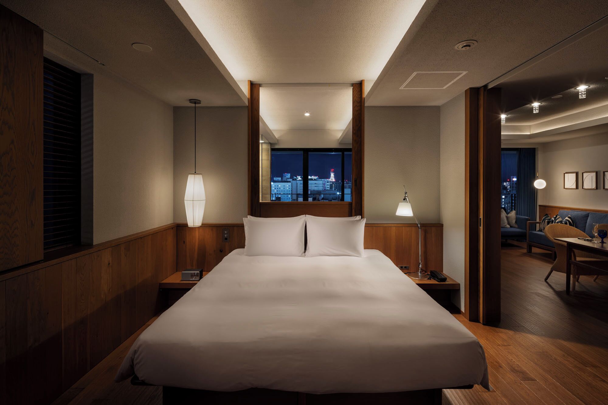 Ein Hotelzimmer mit einem großen Bett, einem Nachttisch, einer Lampe, Blick auf die beleuchtete Stadt und einer Sitzecke mit Tisch und Stühlen.