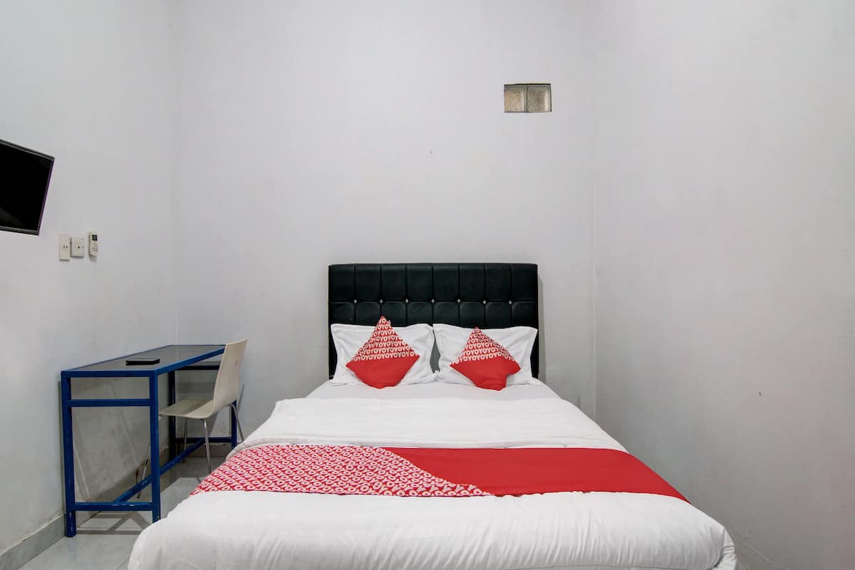 Un lit avec une tête de lit noire, une literie blanche et des coussins rouges. Une petite table avec une chaise se trouve à gauche.
