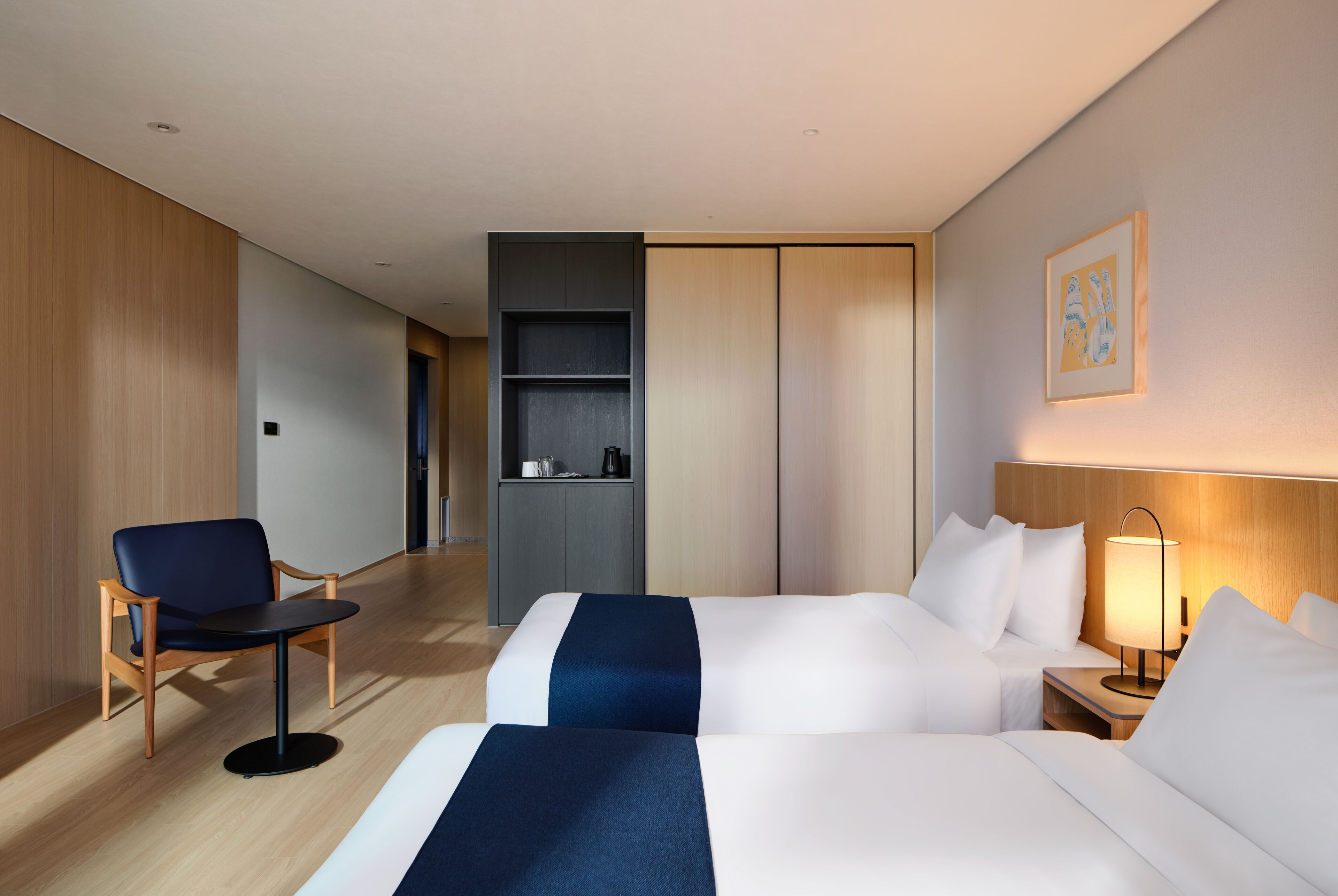 Ein Hotelzimmer mit zwei Betten, einem Schreibtisch, einem Stuhl, einem Kleiderschrank und einem Nachttisch mit Lampe.