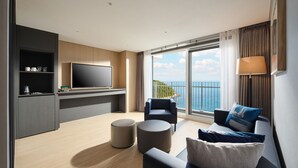 A modern living room with a flat-screen TV, a sofa, two armchairs, a coffee table, and a large window with a view of the sea.