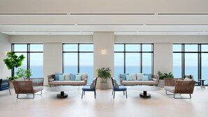 A spacious lounge with a large window offering a view of the sea, a central plant, and a seating area with sofas and chairs.