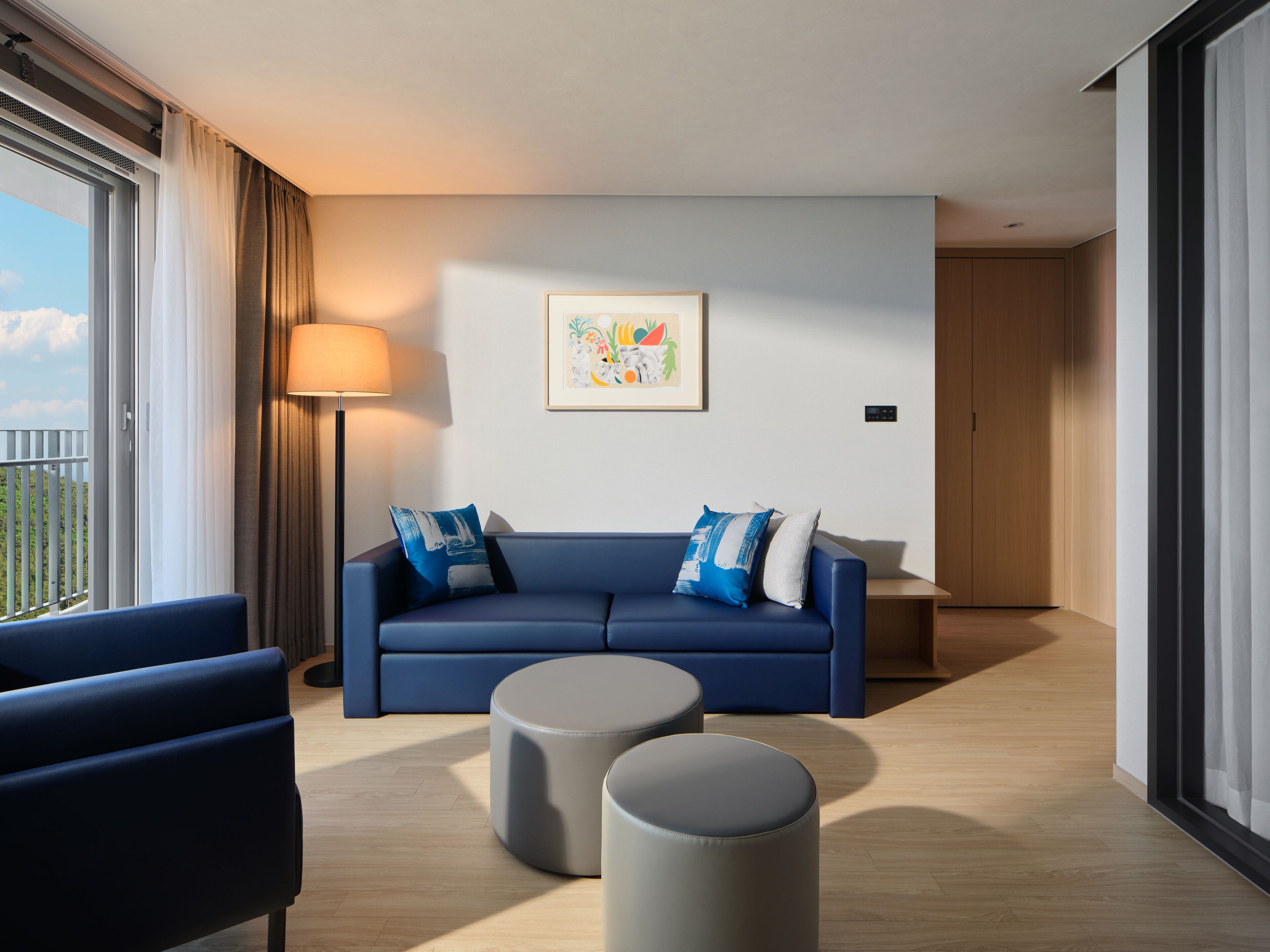 Ein modernes Hotelzimmer mit einem blauen Sofa, zwei Hocker, einer Stehlampe und einem großen Fenster mit Vorhängen.