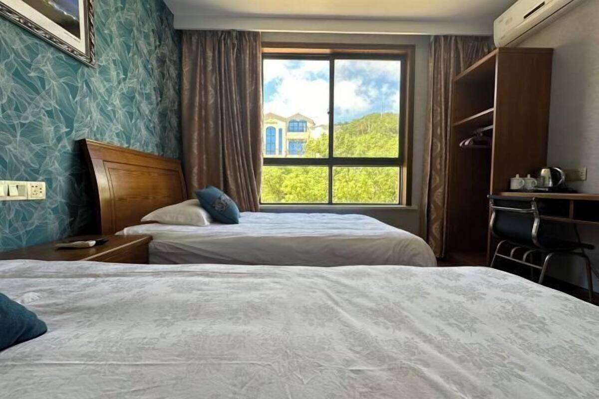 Hotelkamer met twee bedden, een kledingkast, een bureau en een raam met uitzicht op een gebouw en groen.