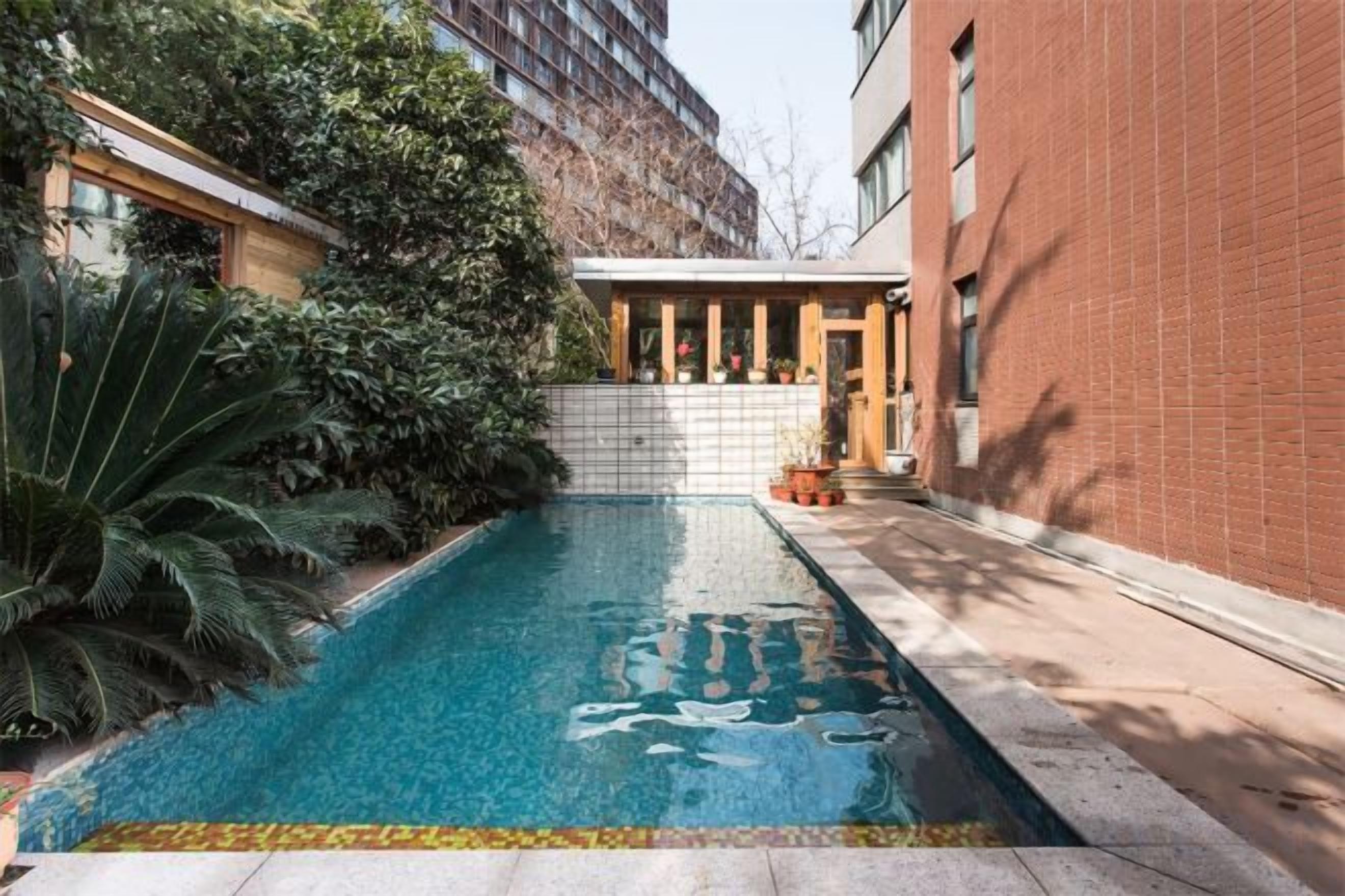 A swimming pool with a tiled border, surrounded by greenery and a brick building.