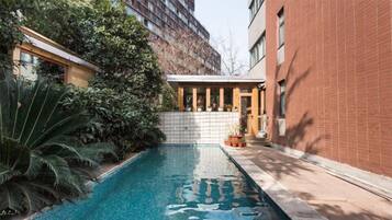 A swimming pool with a tiled border, surrounded by greenery and a brick building.