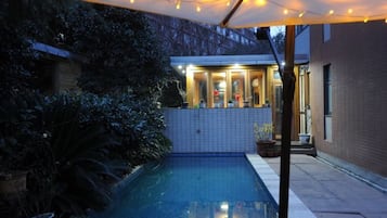 A swimming pool with a tiled deck, surrounded by potted plants and a covered area with string lights.