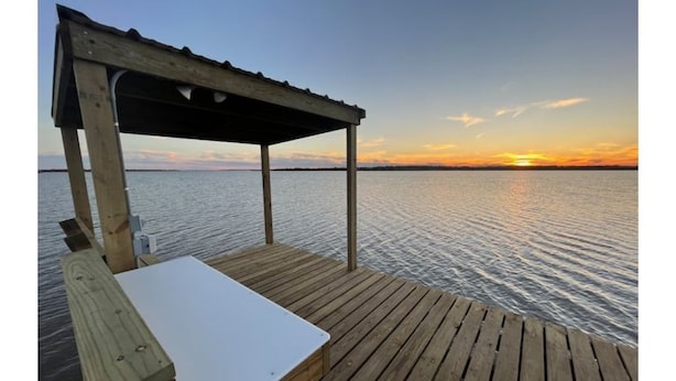 A wooden deck with a table and a covered area overlooking a calm body of water during sunset.