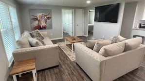 A modern living room with two beige sofas, a coffee table, and a wooden side table. A bull painting adorns the wall, and a flat-screen TV is mounted on the wall.