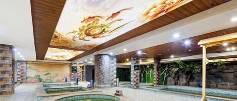 Amplia zona de piscina interior con varias piscinas y un mural decorativo en el techo.