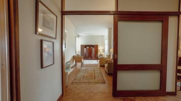 A hallway with wooden flooring, framed artwork on the walls, and a seating area with chairs and a rug.