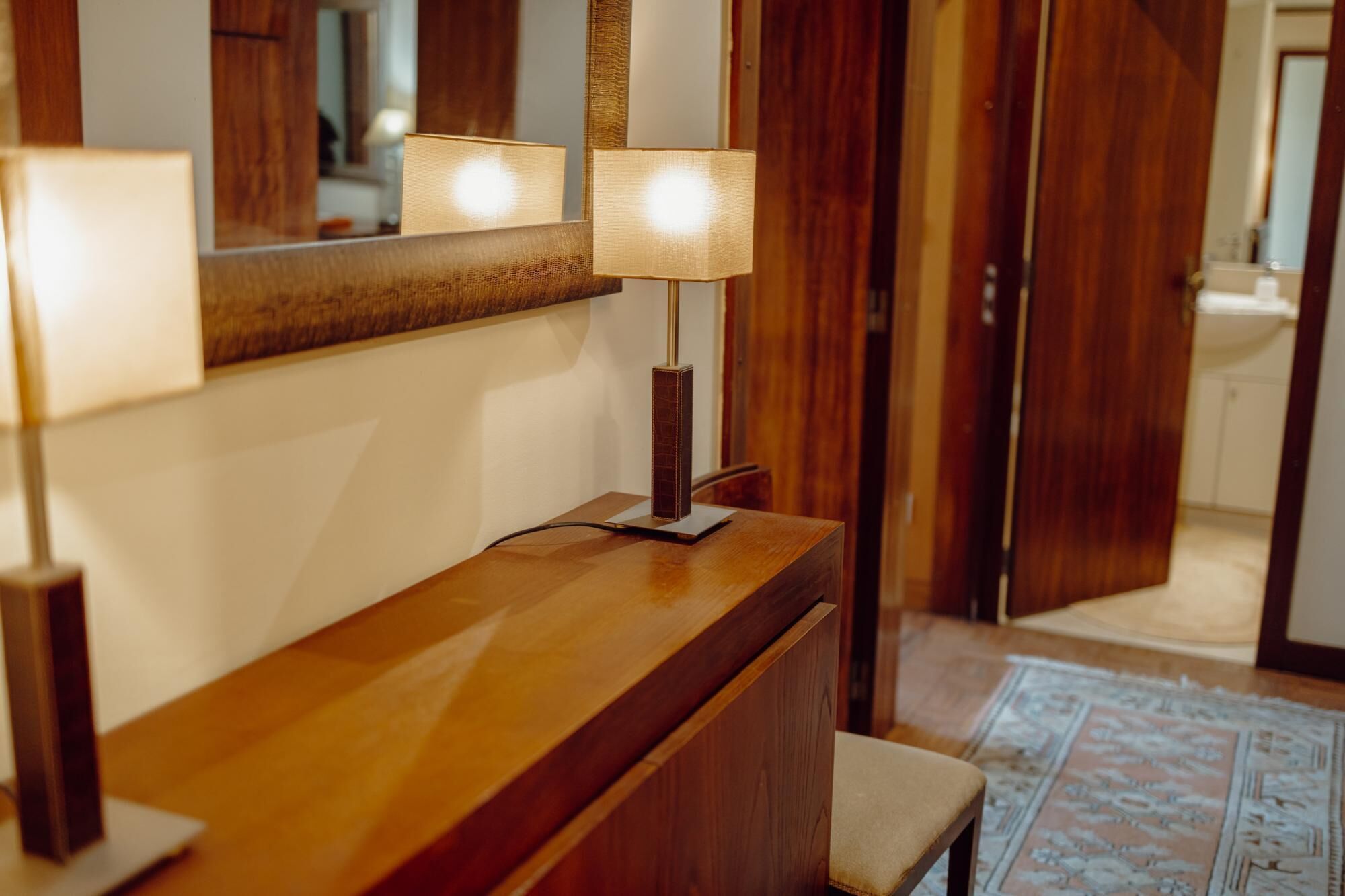 A wooden vanity with a mirror and two lamps. A bench is visible in the foreground.