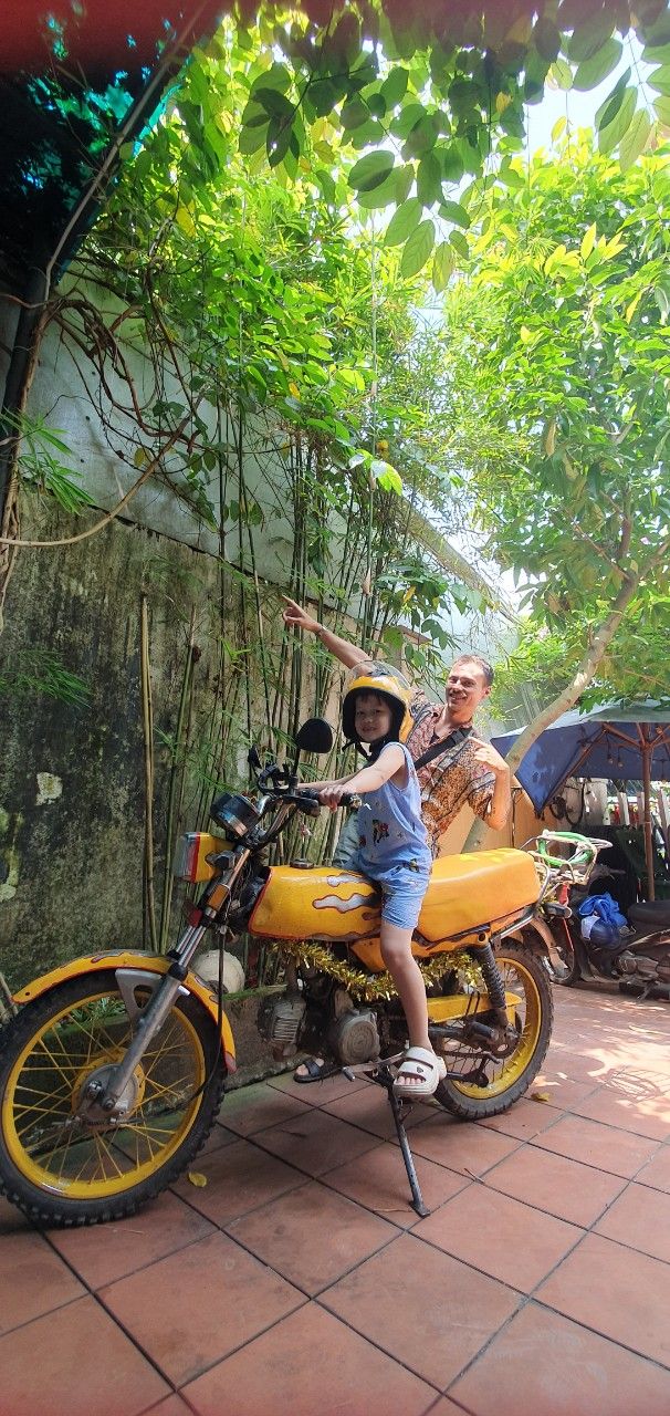 A child sitting on a yellow motorcycle with a helmet, an adult standing behind, and a tiled floor.