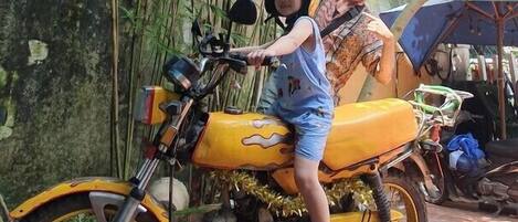 A child sitting on a yellow motorcycle with a helmet, an adult standing behind, and a tiled floor.