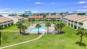 Un resort con piscina central rodeada de palmeras, edificios de varios pisos con balcones y un césped bien cuidado.
