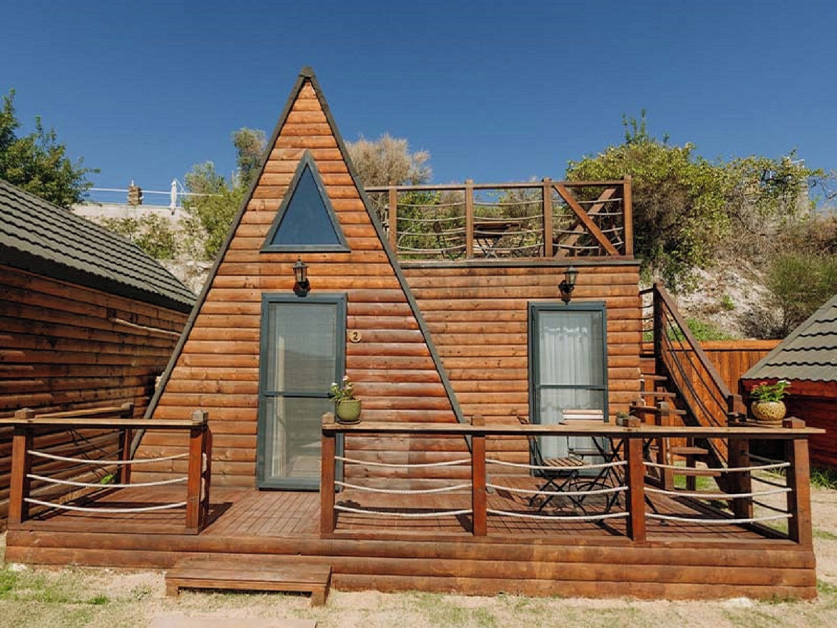 A wooden cabin with a triangular roof, a front door, and a small porch with a table and chairs.
